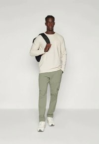 Avslappet antrekk med en lys beige langermet skjorte, olivengrønne cargo bukser og hvite joggesko, samt en svart ryggsekk.