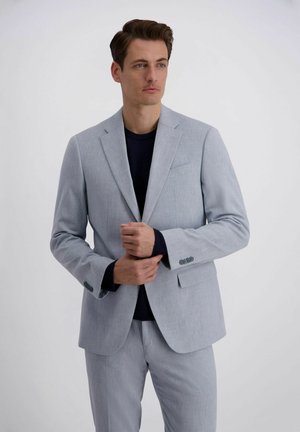 Homme ajustant les manches de sa veste de costume gris clair sur une chemise noire, debout devant un fond blanc uni.