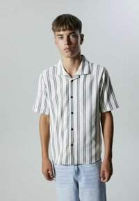 Kortermet skjorte med knapper i hvit med marineblå vertikale striper, med krage, svarte knapper og en avslappet passform. Kombinert med lyse denim.