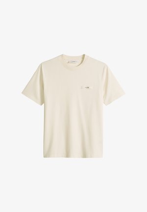 Kreminės spalvos trumpomis rankovėmis suapvalintu kaklu marškinėliai su mažu stačiakampiu lopu kairėje krūtinės pusėje, kuriame yra minimalus juodas tekstas.