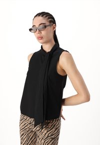 Top sans manches noir avec un détail de décolleté noué, associé à un pantalon rayé beige et marron. Texture de tissu lisse et coupe ajustée.
