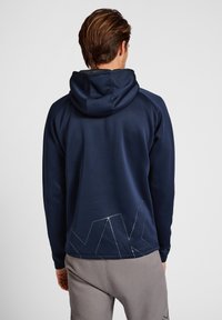 Granatowa bluza z kapturem o luźnym kroju, z centralnym kapturem i geometrycznym logo na dolnej części pleców, wykonana z miękkiego materiału.