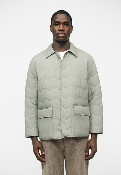 Homme portant une veste matelassée gris clair avec boutons-pression et poches à rabat, debout devant un fond blanc uni.