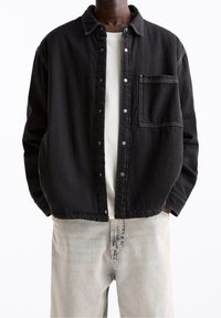 Sort denimjakke med knapper foran, krave og brystlomme. Har kontrastsyninger og en løs pasform. Båret over en hvid skjorte.