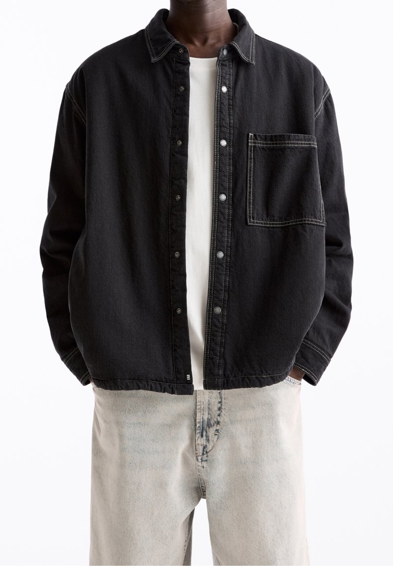 Sort denimjakke med knapper foran, krave og brystlomme. Har kontrastsyninger og en løs pasform. Båret over en hvid skjorte.