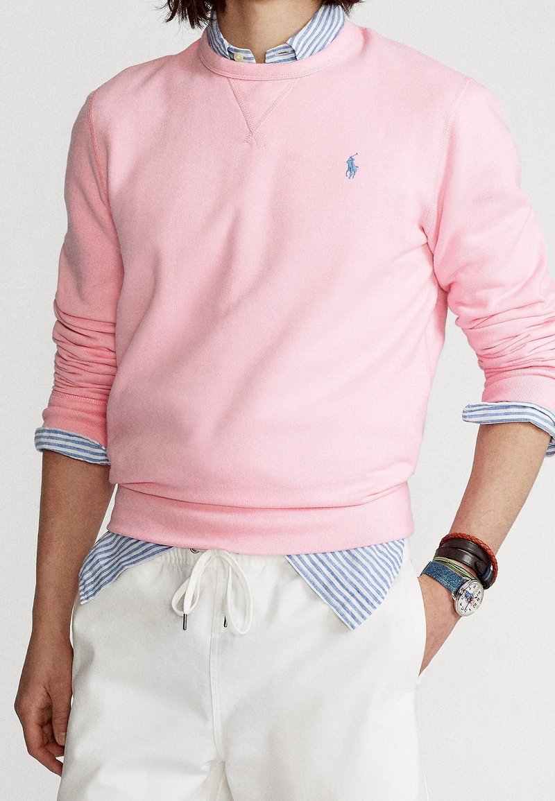 Homme portant un sweat-shirt rose sur une chemise rayée bleue, avec un pantalon blanc à cordon de serrage, une montre et des bracelets au poignet.