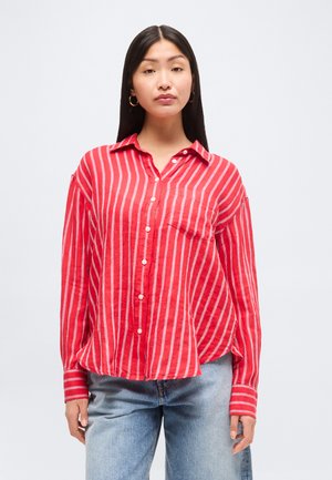 Žena s dlhými čiernymi vlasmi, ktorá nosí zlaté náušnice v tvare kruhu, červeno-bielo pruhovanú košeľu na gombíky a svetlomodré rifle, stojaca pred bielym pozadím.