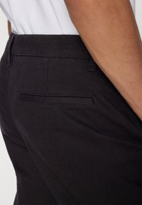 Černé bavlněné kalhoty s hladkým povrchem, obsahující zadní kapsu a štíhlý design. Pás je elastický pro větší pohodlí.