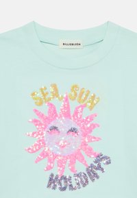 Ανοιχτό μπλε βαμβακερό μπλουζάκι με λαμπερό ροζ σχέδιο ήλιου με παγιέτες, κείμενο που αναγράφει "SEA SUN HOLIDAYS" σε μεταλλικά χρώματα από κάτω.