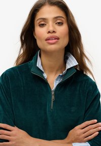 Sudadera de cremallera de terciopelo verde oscuro con un alto cuello, superpuesta sobre una camisa de cuello con rayas azul claro. Presenta un logotipo discreto.