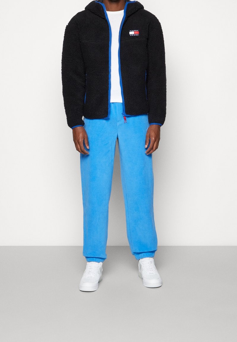 Svart, fluffig zip-up hoodie med blå detaljer; kombinerad med ljusblå fleecebyxor och vita sneakers. En enkel, avslappnad outfit.