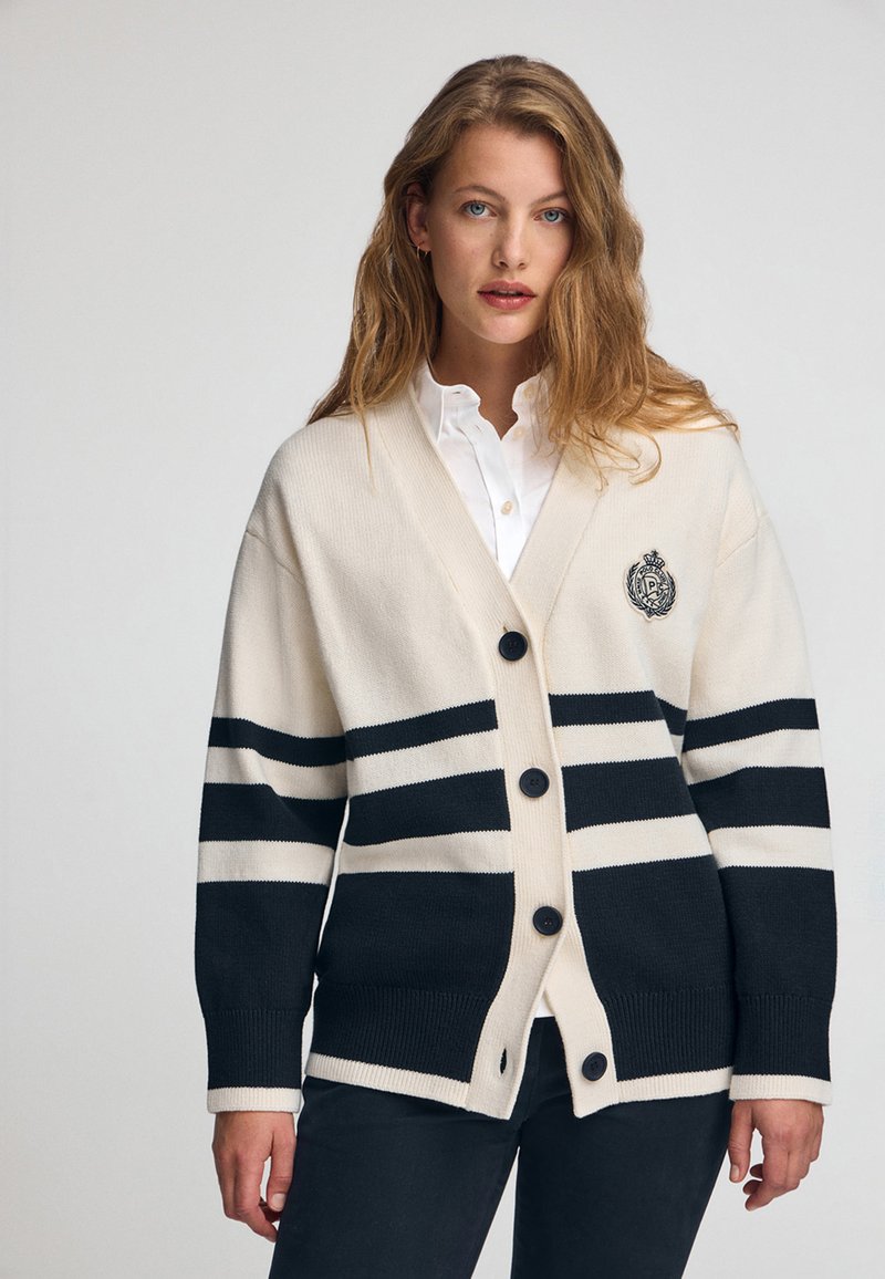 Cremfarbener und marineblauer gestreifter Cardigan mit V-Ausschnitt, Knopfverschluss und gerippten Bündchen. Verfügt über ein Wappen auf der linken Brust.