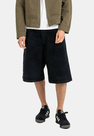 Ein Mann trägt schwarze, breite knielange Cordshorts, schwarze Adidas-Sneaker, eine grüne Jacke und ein weißes Hemd vor einem schlichten Hintergrund.