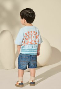 Helles blaues T-Shirt mit kurzen Ärmeln und orangefarbenem Graf_Text "COOL SUNNY DAYS", kombiniert mit aufgerollten Jeansshorts. Beige und blaue Schuhe.