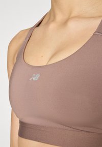Sporta krūšturis mīkstā mauve audumā ar gludu tekstūru, ar noapaļotu kakla izgriezumu un smalku New Balance logo priekšā.