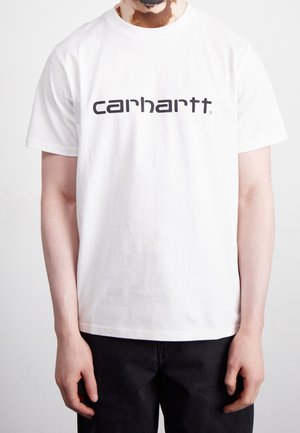 Persoon met vitiligo die een wit t-shirt met Carhartt-logo en een zwarte broek draagt, staand tegen een effen lichte achtergrond.