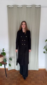 Cappotto nero doppiopetto con bottoni dorati, maniche lunghe e una vestibilità sagomata, abbinato a pantaloni larghi neri e a una piccola borsa nera.