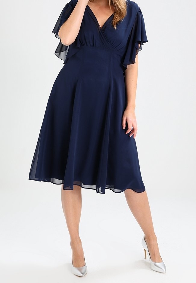 Robe en chiffon bleu marine avec des manches flottantes et un décolleté en V. Silhouette évasée en A, longueur genou, associée à des talons hauts argentés.