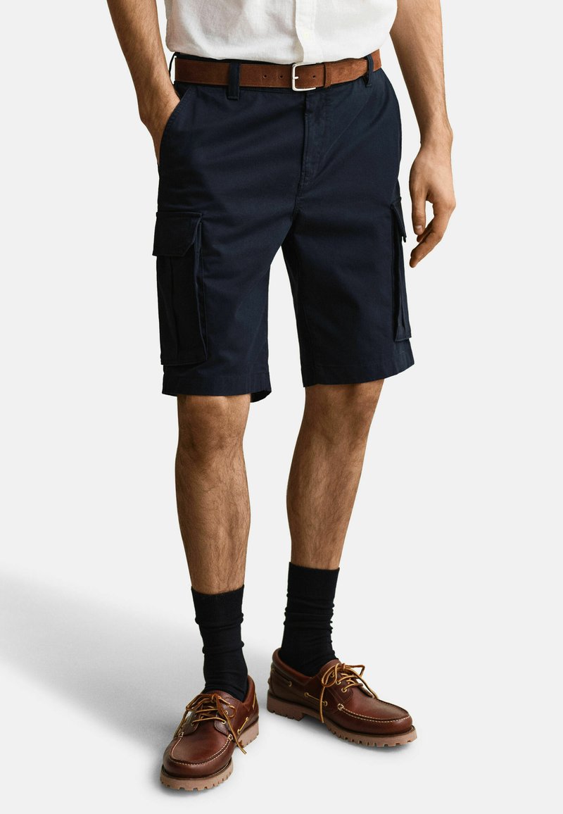 Homme portant un short cargo bleu marine, une ceinture en cuir marron, des chaussettes noires et des chaussures à lacets en cuir marron, debout les mains détendues le long du corps.