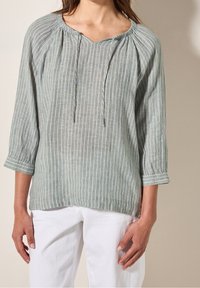 Femme portant un chemisier à rayures verticales bleues claires et blanches avec des manches 3/4 et un pantalon blanc, debout contre un mur beige.