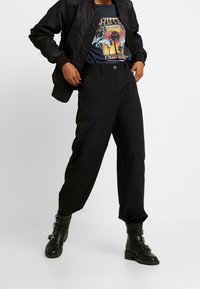 Schwarze Bomberjacke über einem T-Shirt mit grafischem, buntem Print, kombiniert mit locker sitzenden schwarzen Hosen und kniehohen schwarzen Stiefeln mit Schnallen.