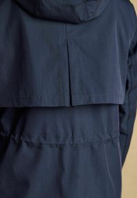 Giacca impermeabile blu scuro con cappuccio, caratterizzata da un design posteriore a strati e vita cinchata. Texture morbida e dettagli di cucitura sottili.