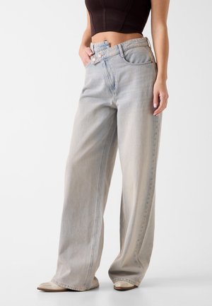 Vrouw draagt lichtblauwe wijd uitlopende jeans met dubbele knoopsluiting en een zwart kort topje, staat met hand in zak.