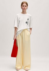 Witte oversized t-shirt met een zwart vaas en plant grafiek, gecombineerd met gele wijde broek. Model houdt een rode tote bag vast.