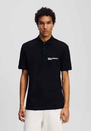 Schwarzes Poloshirt aus Baumwolle, mit Kragen, drei Knöpfen und einem weißen Logo auf der linken Brustseite. Kurzärmlig, unifarben.