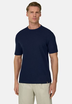Homme portant un t-shirt à manches courtes bleu marine uni avec un col rond et un pantalon beige clair, debout devant un fond blanc.
