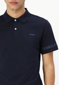 Cămașă polo bleumarin din bumbac; dispune de guler, două butoane și logo-ul "GUESS" brodat pe piept și pe mânecă.