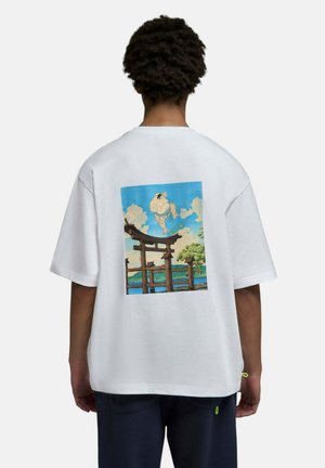 Personne portant un t-shirt blanc avec un graphique d’un lutteur de sumo faisant du skateboard au-dessus d’une porte torii japonaise traditionnelle sur fond de ciel bleu.