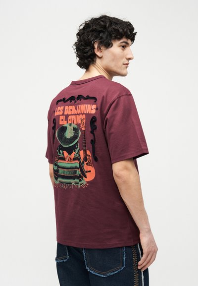 Άνδρας με σγουρά μαλλιά φοράει μπορντό μπλουζάκι με γραφικό μιας φιγούρας με καπέλο, κουβέρτα και κιθάρα, με το κείμενο "Les Benjamins El Gringo" στην πλάτη.