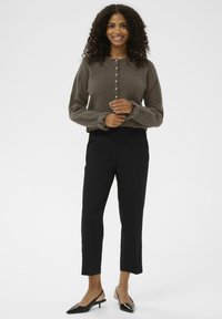 Ženska s kodrasto pričesko se smeška v rjavem puloverju na gumbe, črnih skrajšanih hlačah in črnih elegantnih čevljih s koničastim nosom na belem ozadju.