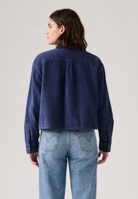 Marineblauwe ribstof shirt met een cropped design, voorzien van een geknoopte manchet en een rugplooi, gecombineerd met lichtblauwe denim jeans.
