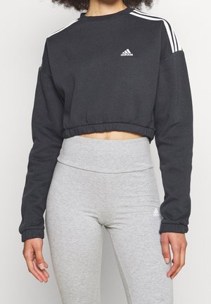 Osoba ubrana w czarną krótką bluzę Adidas z białymi paskami oraz szare legginsy Adidas z wysokim stanem, pokazana od szyi do bioder.