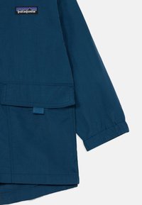Veste Patagonia bleu marine avec un logo sur la poitrine, une poche frontale et des poignets élastiques. Fabriquée en tissu léger avec une texture lisse.