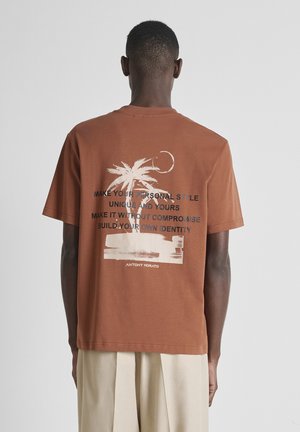 Čovjek u smeđoj majici s bijelom palmom i motivacijskim tekstom stoji okrenut leđima na jednobojnoj pozadini.