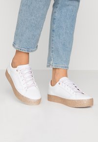 White sneakers with a textured, glittery beige sole. Features include lace-up design and light pink accents on the eyelets and laces.