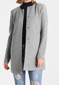 Manteau gris en mélange de laine avec un col rond, une fermeture à pression devant et des manches longues. Tissu texturé avec une coupe droite. Porté par-dessus un jean.