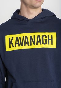 Námořní modrý mikina s velkým žlutým obdélníkovým grafikou, která obsahuje tučný černý text "KAVANAGH." Materiál vypadá měkký a má kapcovou konstrukci.