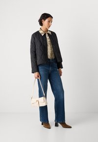 Giacca nera trapuntata, camicia crema a fantasia, jeans blu a gamba larga, borsa beige con accessori dorati e stivaletti alla caviglia in camoscio marrone.