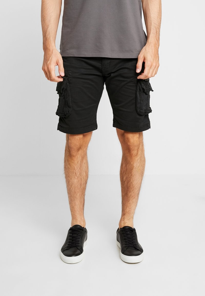Homme portant un short cargo noir, des baskets noires avec des semelles blanches et un t-shirt gris, debout devant un fond clair.
