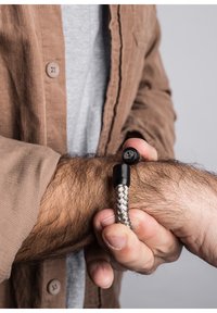 Bracciale in corda intrecciata d'argento con una chiusura nera che presenta un design di loto, indossato su un polso con peli visibili. Sfondo neutro.