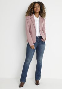 Blazer rose clair avec deux boutons, associé à une chemise blanche à col et un jean évasé bleu. Des bottes à talons brunes complètent la tenue.