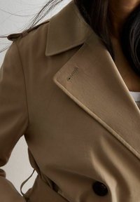 Béžový trenčkot se širokým límcem, dvojím zapínáním a tmavými knoflíky. Hladká látka s jemným leskem a viditelnými švy.