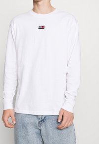 Ein Mann trägt ein weißes Langarmshirt mit einem kleinen roten, weißen und schwarzen Tommy-Logo auf der Brust, kombiniert mit hellblauen Jeans.