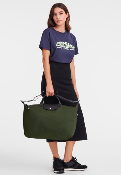 Green nylon tote bag with a black top handle and accents. Features a zipper closure and a spacious design. Model pairs with a navy graphic tee.