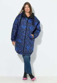 Gequiltede blauwe luipaardprint pufferjas met een zwarte capuchon, ritsluiting en zijzakken. Gedragen over blauwe spijkerbroeken en laarzen met een roze zool.