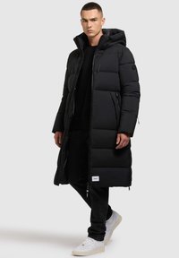 Abrigo largo acolchado negro con capucha, que presenta una cremallera, bolsillos laterales y un parche con logo. Llevado sobre una camiseta negra y jeans negros, con zapatillas blancas.